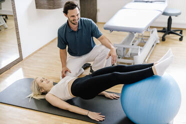 Physiotherapeutin hilft einer Patientin, die mit einem Fitnessball übt, auf einer Gymnastikmatte liegend - DAWF01578