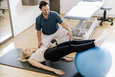 Physiotherapist assisting female patient, practicing with fitness ball, lying on gym mat - DAWF01578