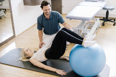 Physiotherapeutin hilft einer Patientin, die mit einem Fitnessball übt, auf einer Gymnastikmatte liegend - DAWF01579