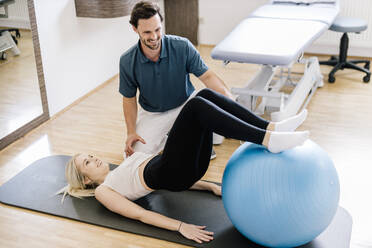 Physiotherapist assisting female patient, practicing with fitness ball, lying on gym mat - DAWF01579