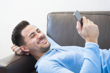 Glücklicher junger Mann, der sein Smartphone benutzt, während er zu Hause auf dem Sofa liegt - DIGF12154