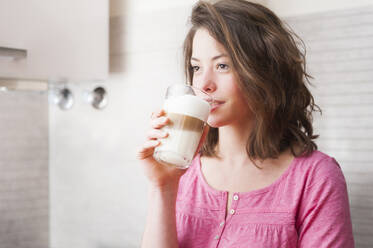 Thoughtful woman drinking Dalgona coffee while standing in kitchen at home - DIGF12155