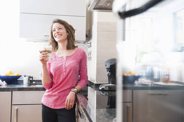 Glückliche Frau hält ein Glas Dalgona-Kaffee in der Hand, während sie zu Hause in der Küche steht - DIGF12156