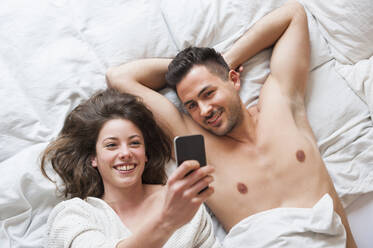 Fröhliche Frau, die ein Selfie mit einem Mann macht, während sie zu Hause im Bett liegt - DIGF12162