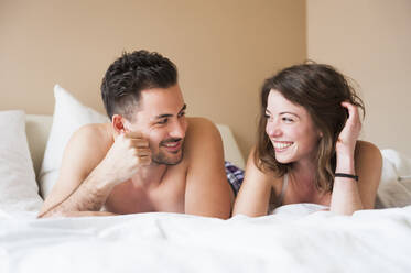 Happy young couple looking at each other while lying in bed at home - DIGF12164