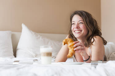 Glückliche junge Frau mit Croissant in der Hand beim Frühstück im Bett - DIGF12165