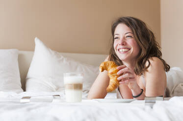 Happy young woman holding croissant while having breakfast in bed - DIGF12165