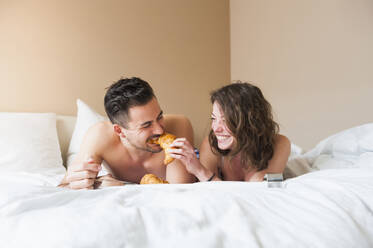 Glückliche junge Frau füttert einen Mann mit einem Croissant, während sie zu Hause im Bett liegt - DIGF12166