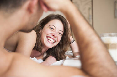 Fröhliche junge Frau, die einen Mann ansieht, während sie zu Hause im Bett liegt - DIGF12169