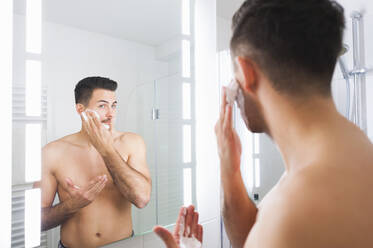 Reflection of young man applying shaving cream on cheek at home - DIGF12181