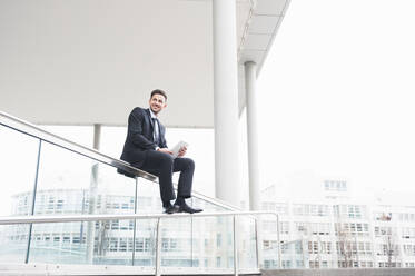 Businessman holding digital tablet while sitting on glass railing - DIGF12183