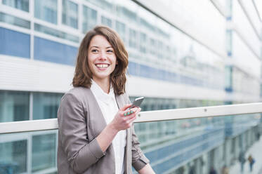 Glückliche Geschäftsfrau hält ihr Smartphone in der Hand, während sie vor einem Glasgebäude steht - DIGF12185