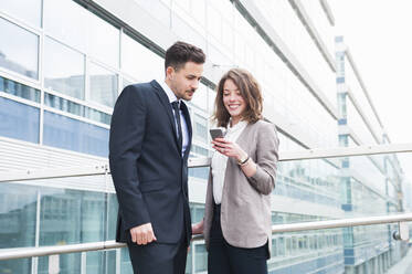 Happy business couple using smart phone while standing outside office building - DIGF12186