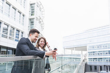 Business couple using smart phone on elevated walkway outside office building - DIGF12188