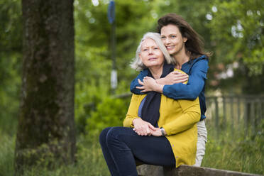 Portrait of happy woman hugging her mother sitting on backrest of a park bench - DIGF12200