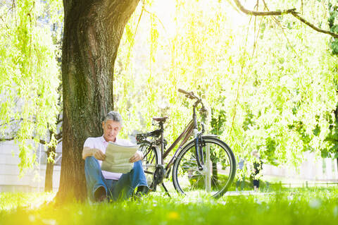 Senior man leaning against tree trunk in a park reading newspaper stock photo