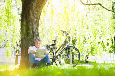 Älterer Mann lehnt an einem Baumstamm in einem Park und liest Zeitung - DIGF12253