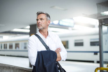 Thoughtful businessman with arms crossed looking away while standing at railroad station platform - DIGF12276
