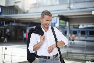 Businessman holding blazer checking time while standing against railroad station - DIGF12278
