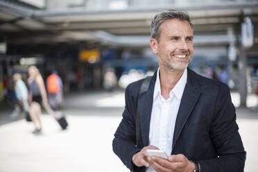 Lächelnder Geschäftsmann, der ein Smartphone hält und wegschaut, während er am Bahnhof steht - DIGF12282