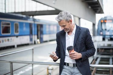 Geschäftsmann mit Kaffee in der Hand und Smartphone auf dem Bahnhof - DIGF12283