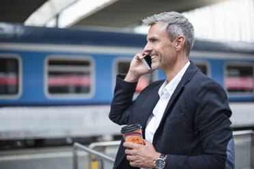 Geschäftsmann mit Kaffee in der Hand, der über sein Smartphone spricht, während er am Bahnhof steht - DIGF12284