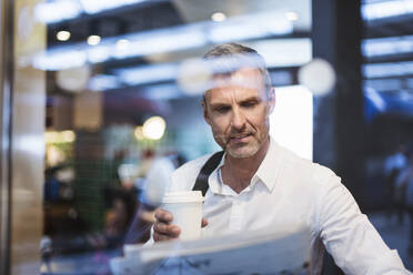 Businessman holding coffee reading newspaper while sitting in train seen through window - DIGF12288