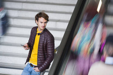 Porträt eines jungen Mannes mit Smartphone, der an einer Treppe wartet - DIGF12296