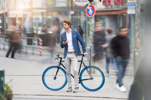 Porträt eines jungen Geschäftsmannes mit Fahrrad und Smartphone, der in der Stadt wartet - DIGF12299
