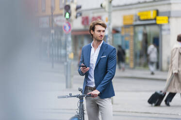 Porträt eines jungen Geschäftsmannes mit Fahrrad und Smartphone, der in der Stadt wartet - DIGF12300