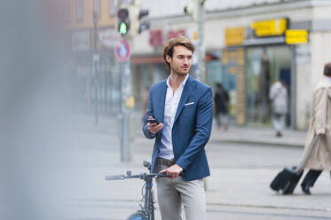 Portrait of young businessman with bicycle and smartphone waiting in the city - DIGF12300