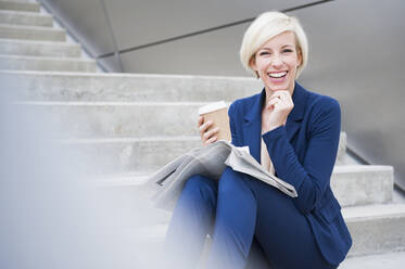 Portrait of blond laughing businesswoman with coffee to go and newspaper sitting on stairs - DIGF12362