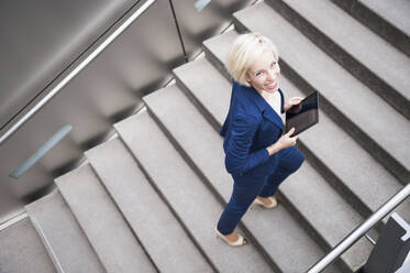 Porträt einer lächelnden blonden Geschäftsfrau beim Treppensteigen mit Tablet - DIGF12363
