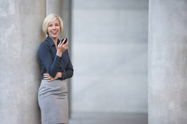 Lachende blonde Geschäftsfrau mit Smartphone an Säule stehend - DIGF12370
