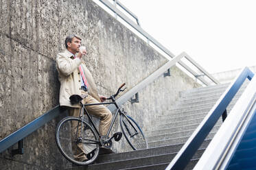 Businessman drinking coffee while standing with bicycle on steps - DIGF12372