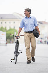 Lächelnder Geschäftsmann schaut weg, während er mit dem Fahrrad auf der Straße in der Stadt spazieren geht - DIGF12377