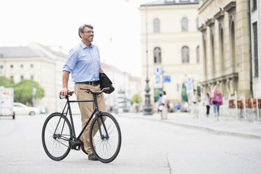 Smiling businessman looking away while walking with bicycle on road in city - DIGF12378