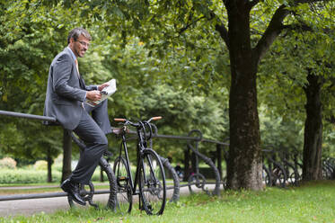 Businessman reading newspaper while sitting on railing in public park - DIGF12379