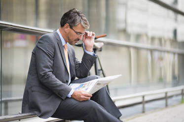 Businessman wearing eyeglasses reading newspaper while sitting on railing - DIGF12381