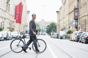 Businessman wearing suit with bicycle crossing road in city - DIGF12384