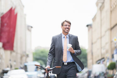 Smiling businessman holding coffee looking away while walking with bicycle in city - DIGF12385