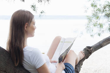 Smiling woman sitting on tree trunk with magazine at Ammersee looking at distance, Germany - DIGF12408