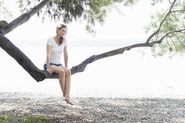 Lächelnde Frau sitzt auf einem Baumstamm am Ammersee und entspannt sich, Deutschland - DIGF12410