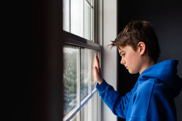 Tween boy looking out of a window in a dark room. - CAVF83055
