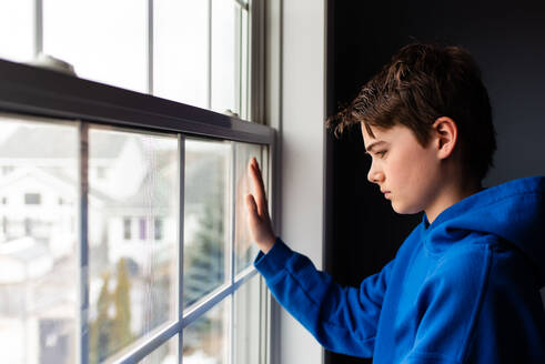 Kleiner Junge, der in einem dunklen Raum aus dem Fenster schaut. - CAVF83056