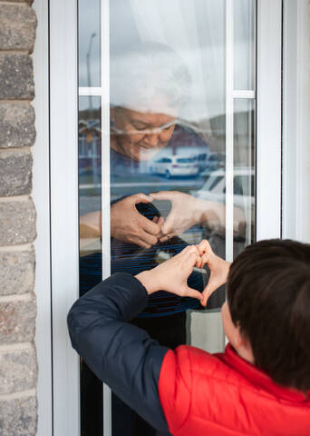 Kleiner Junge schaut während der Pandemie Covid 19 durch das Fenster auf seine Großmutter., lizenzfreies Stockfoto