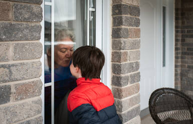Kleiner Junge schaut während der Pandemie Covid 19 durch das Fenster auf seine Großmutter. - CAVF83071