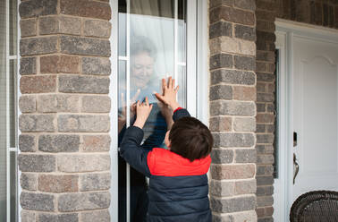Kleiner Junge schaut während der Pandemie Covid 19 durch das Fenster auf seine Großmutter. - CAVF83072