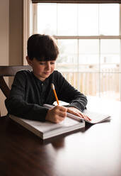 Junge macht Schularbeiten in einem Arbeitsbuch zu Hause am Tisch. - CAVF83082