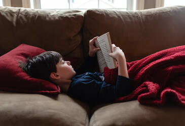 Kleiner Junge liest auf einem Sofa unter einer Decke ein Buch. - CAVF83086